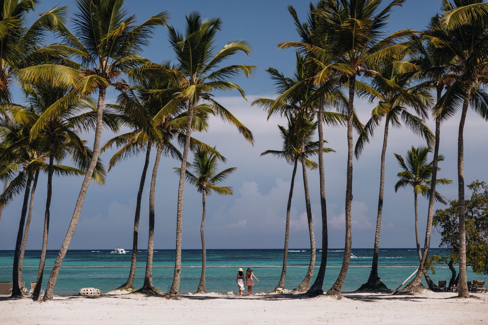 Two women walk along a sandy beach lined with palm trees, overlooking the clear blue ocean at St. Regis Cap Cana Resort.