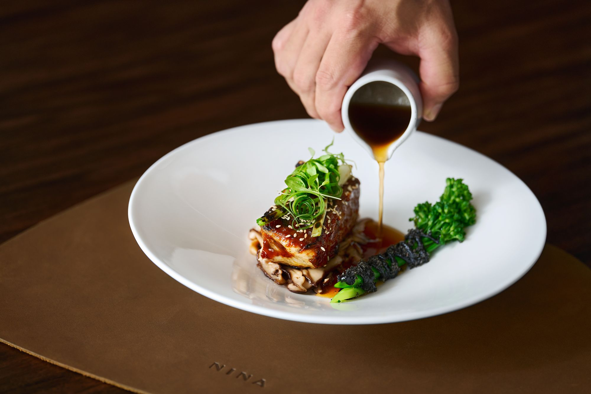 A hand pours sauce over a beautifully plated dish featuring grilled fish, garnished with green onions and fresh vegetables.