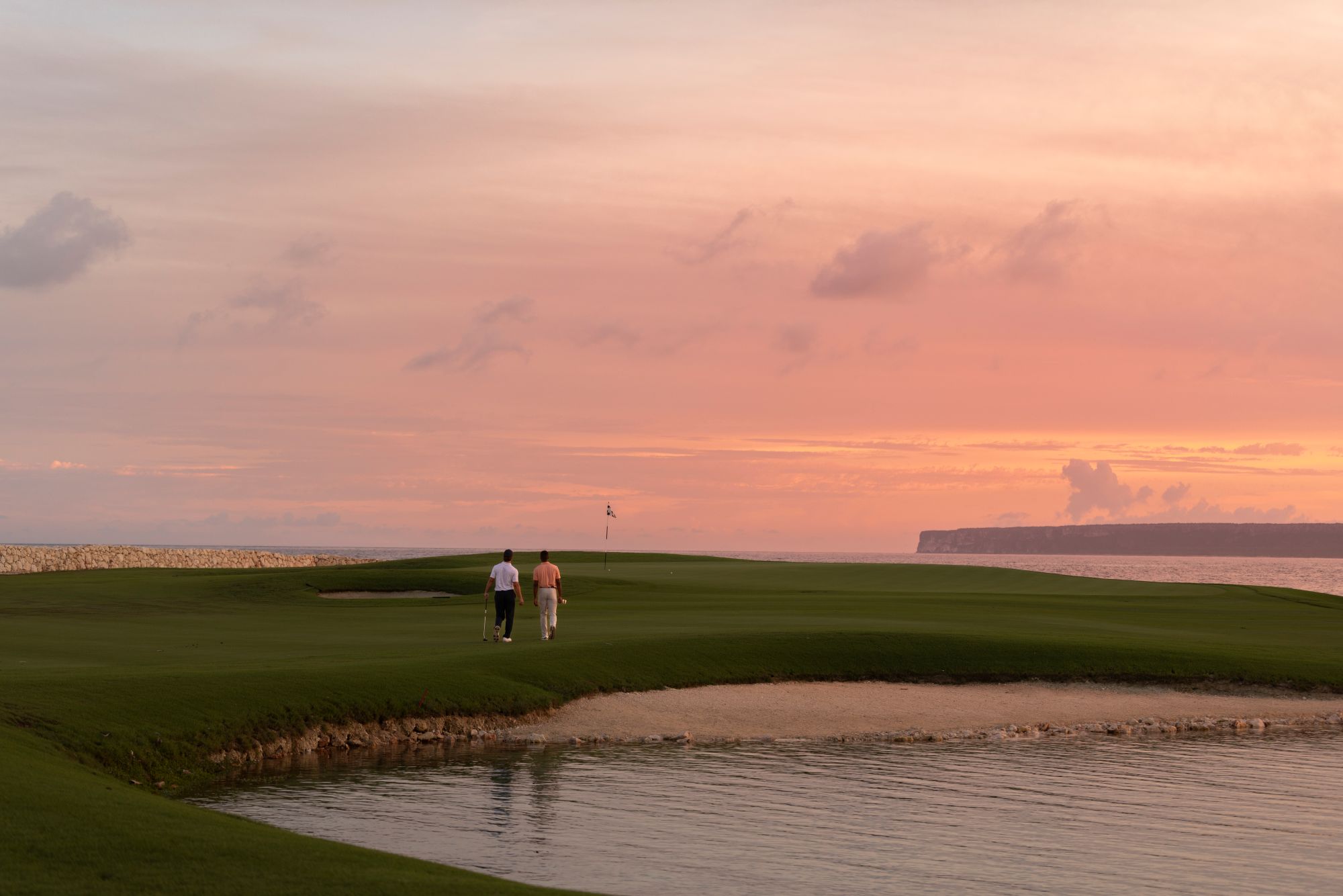 Two golfers walk along a lush green golf course at sunset, with water and cliffs visible in the background.