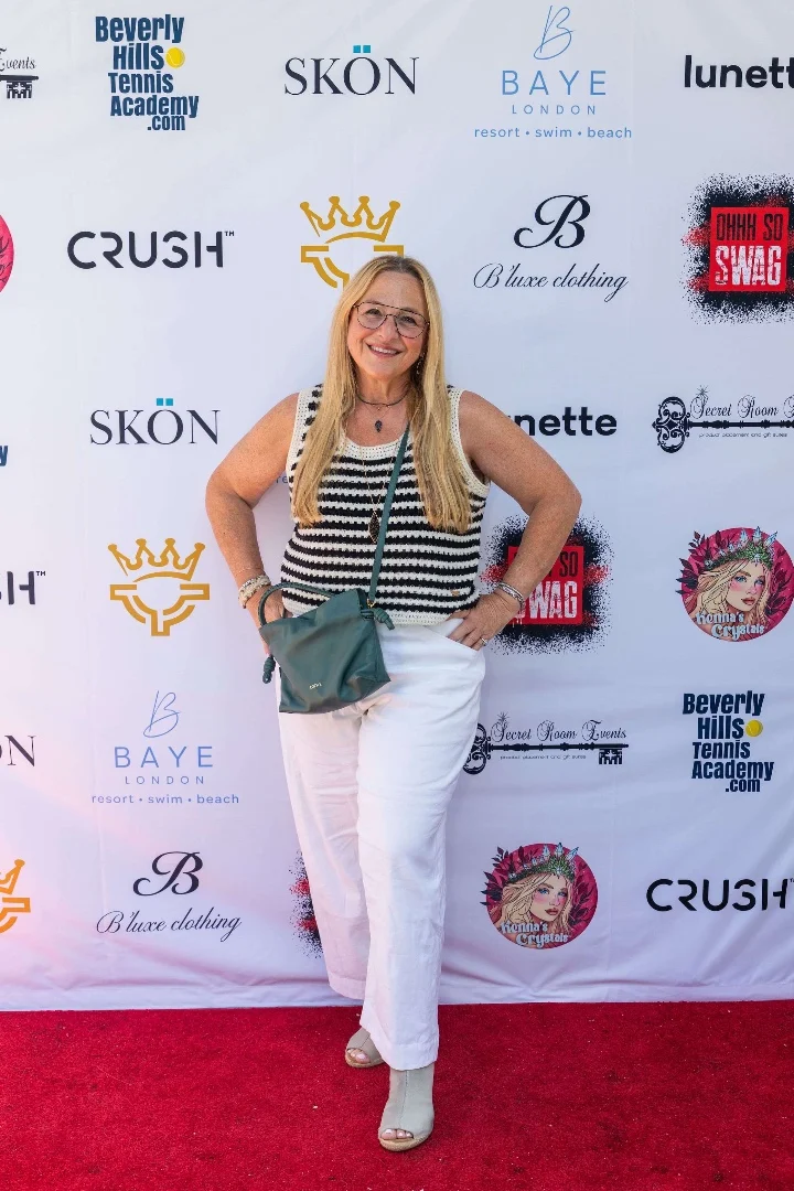 Smiling woman in a striped top and white pants posing on a red carpet with event sponsor logos in the background.