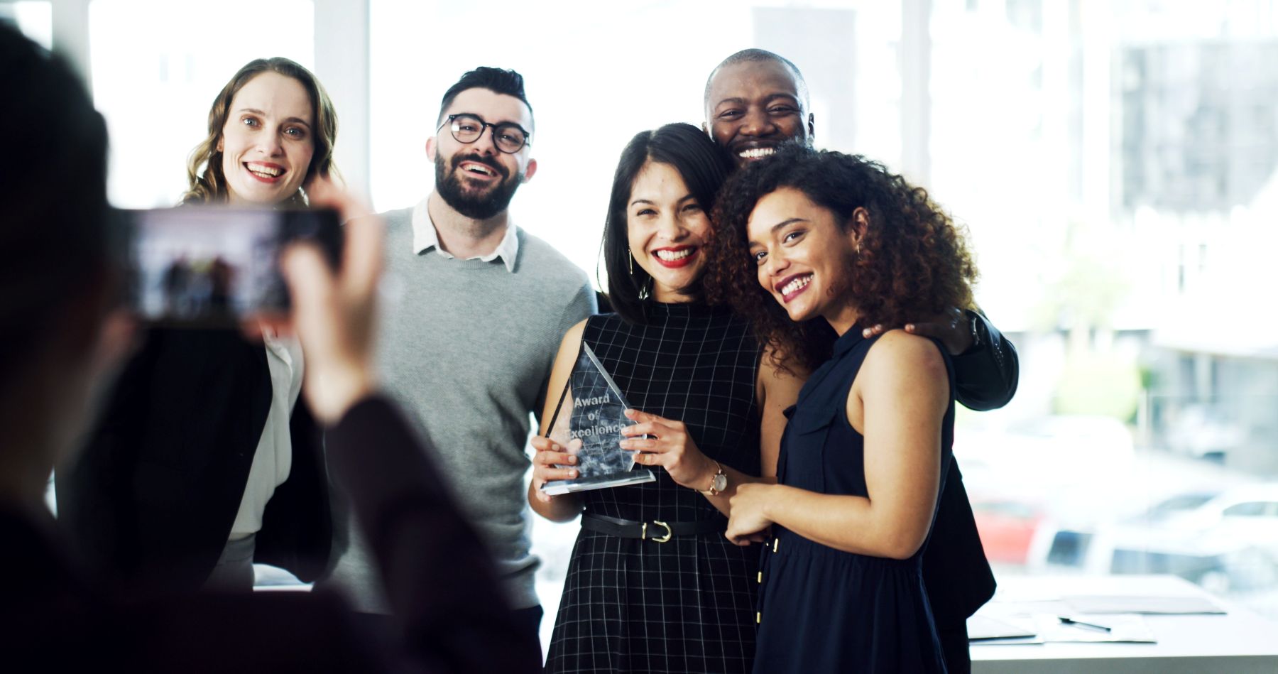 A group of five smiling people celebrate as one holds an award for excellence in a bright office setting.