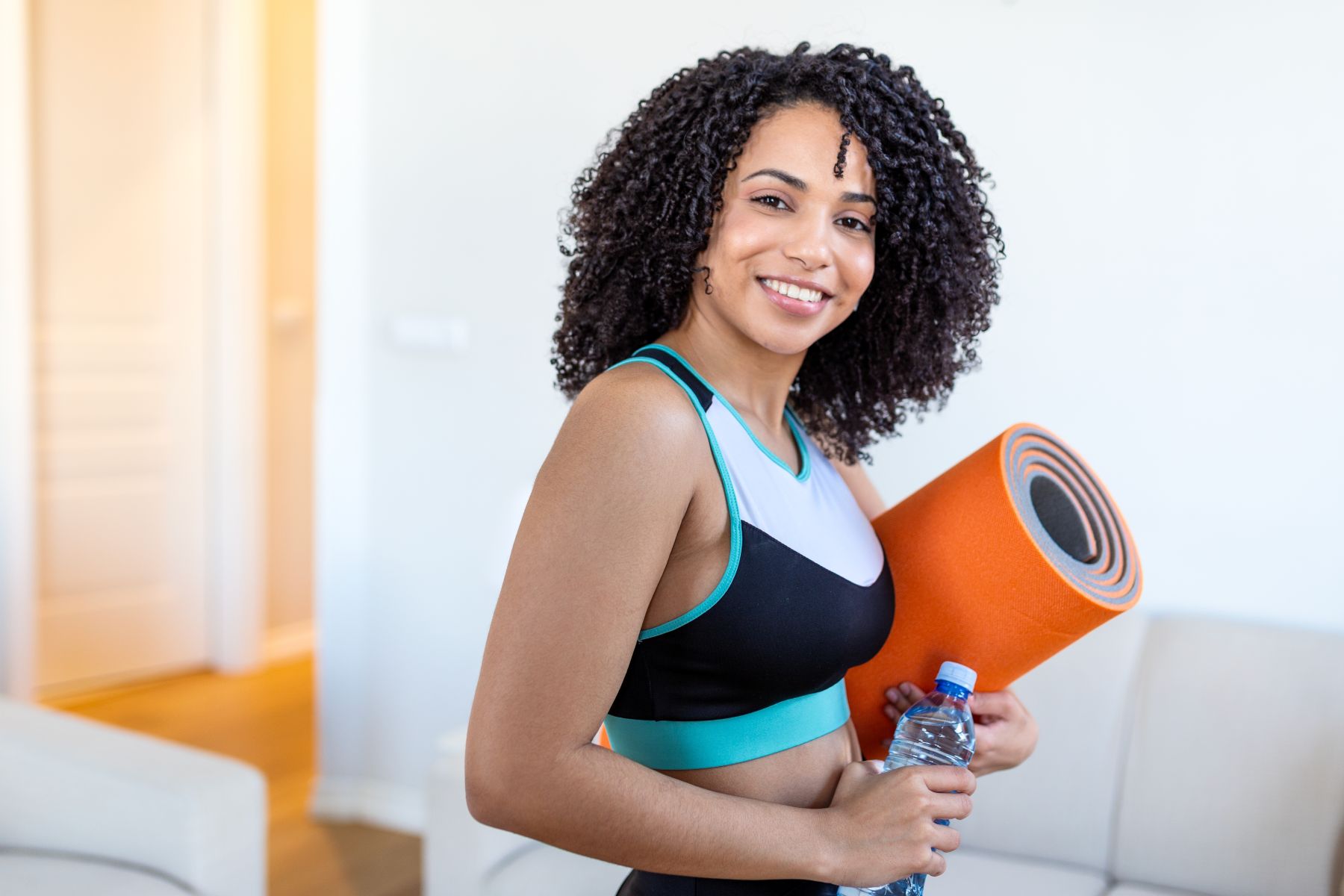 Smiling woman holding a water bottle and yoga mat, ready for her first Pilates class in a bright indoor space.