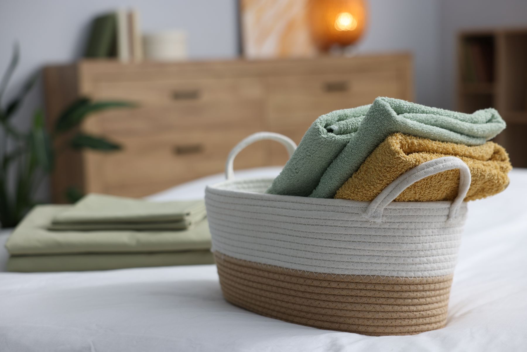 A cozy basket holds luxurious bath sheets in green and yellow, set against a backdrop of a serene bedroom environment.