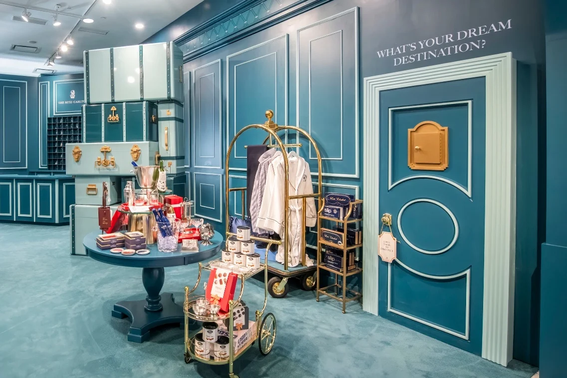 Interior of a Bloomingdale's display featuring The Ritz-Carlton, with blue walls, luggage, a table with products, and a stylish cart.