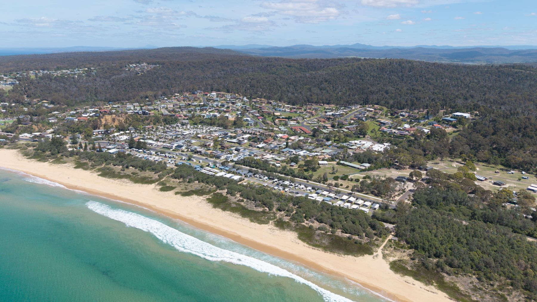 Aerial view of a coastal town with residential areas, beach, and greenery, highlighting the serene environment for visitors.