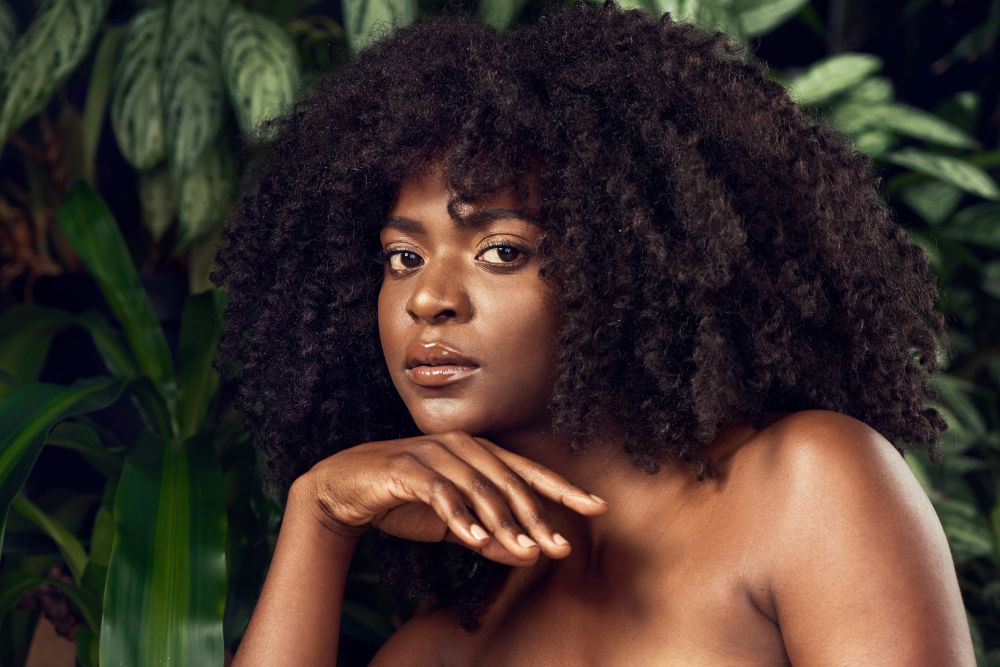 A woman with natural curly hair poses thoughtfully against lush green foliage, embodying high fashion and beauty.