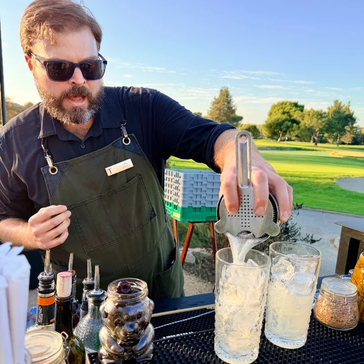 Bartender straining a cocktail mixture into two glasses filled with ice at a live-fire dinner event in the chef's garden.