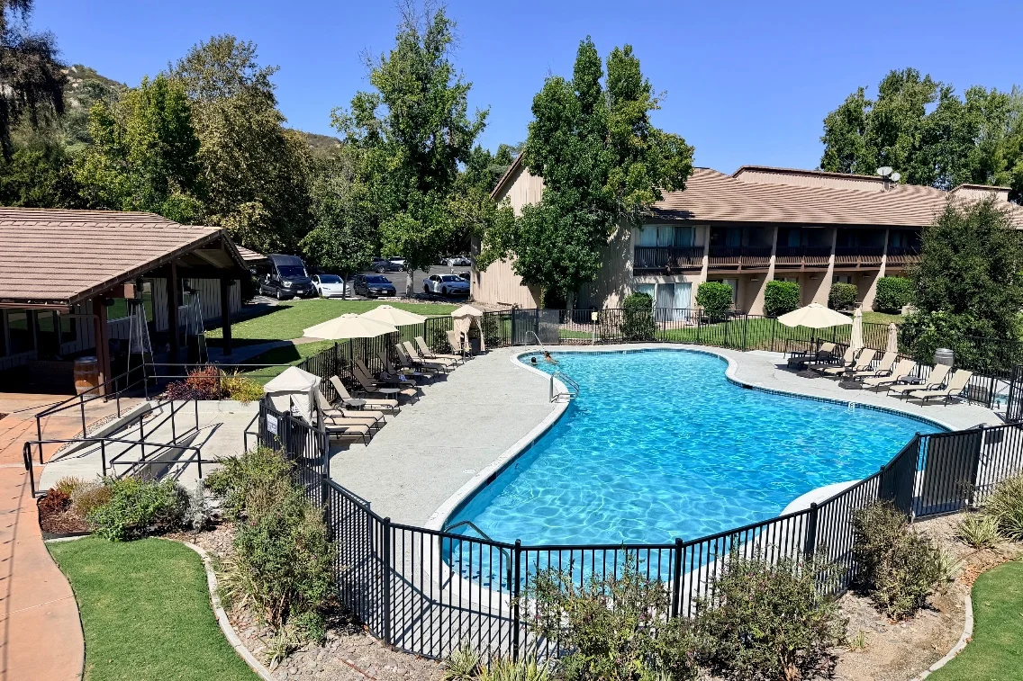 A clear swimming pool surrounded by lounge chairs and umbrellas, with a building and trees in the background at Temecula Creek Inn.