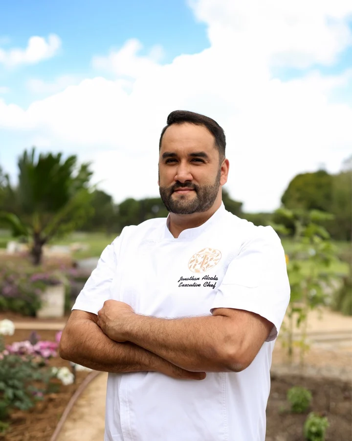 Executive chef Jonathan Alcala poses confidently in a garden at Temecula Creek Inn, surrounded by lush greenery.