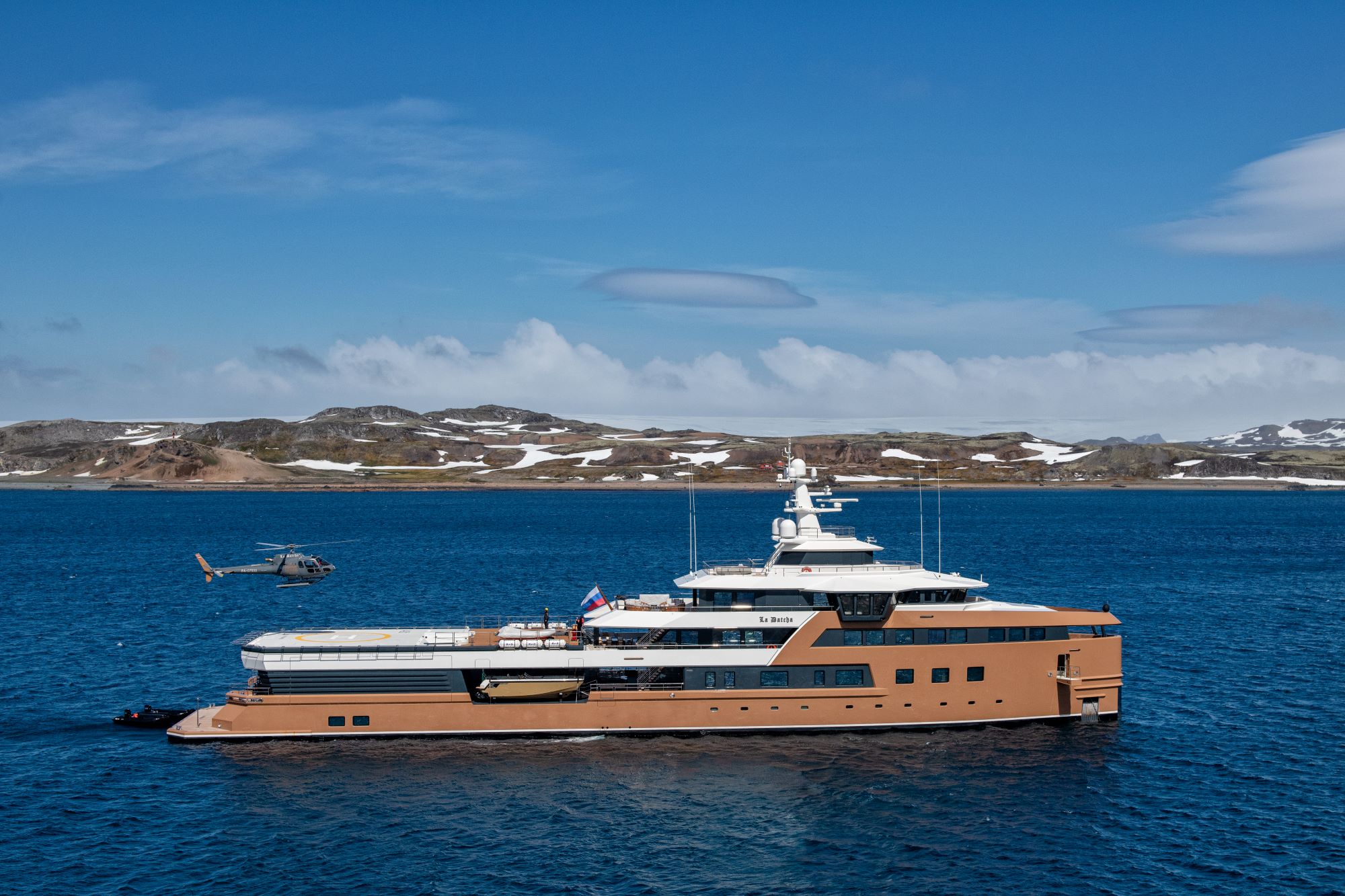 LA Datcha luxury yacht in clear blue water, with a helicopter flying above and rugged coastline in the background.