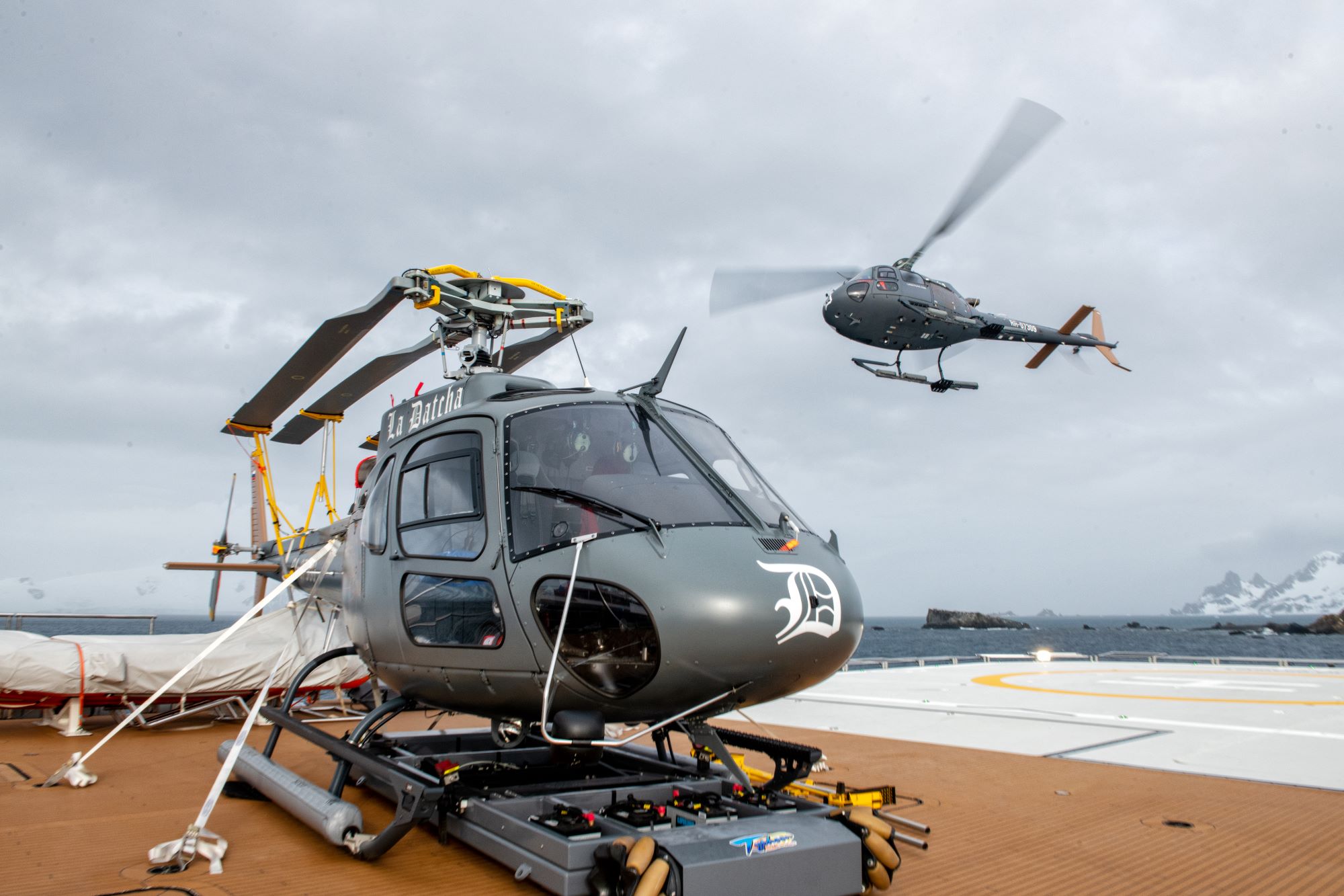 Helicopters operated by La Datcha on a yacht's helipad, with rugged landscape and ocean views in the background.