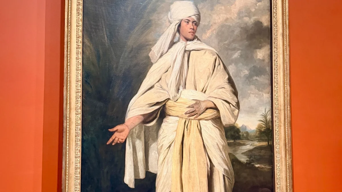 Portrait of a young man dressed in traditional attire, gesturing with his right hand against a dramatic background.