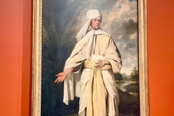 Portrait of a young man dressed in traditional attire, gesturing with his right hand against a dramatic background.