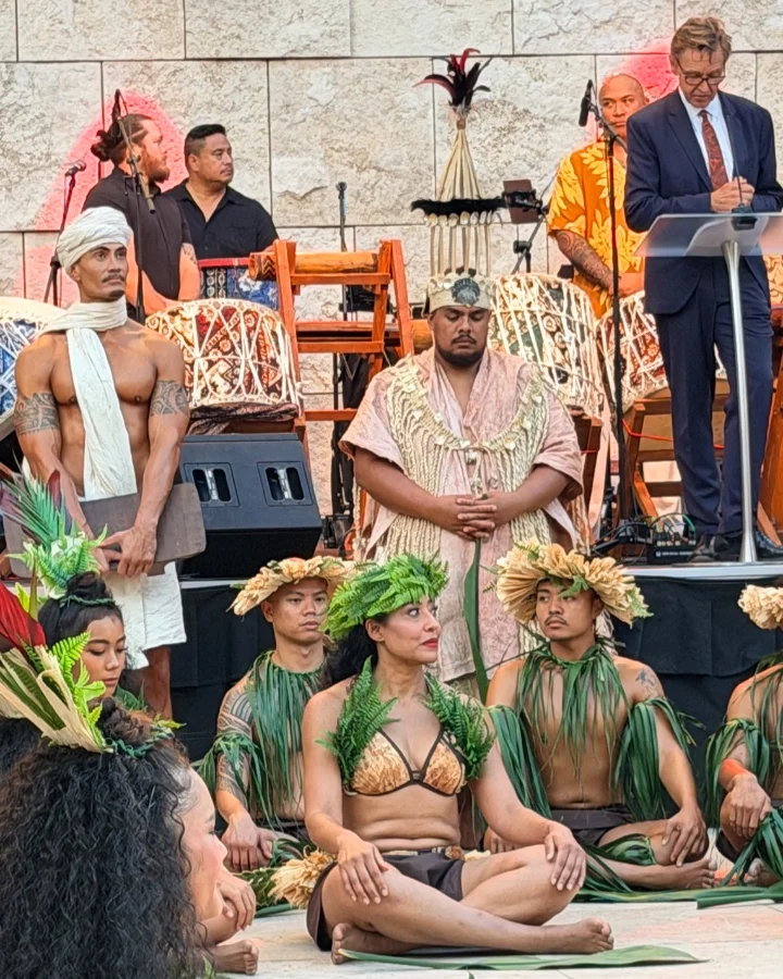 Ceremonial presentation with performers in traditional attire, featuring men and women seated with greenery, as a speaker addresses the audience.
