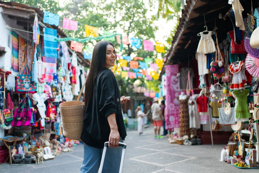 Traveler with a suitcase and woven bag smiles while exploring a vibrant market in Mexico, surrounded by colorful stalls and decorations.