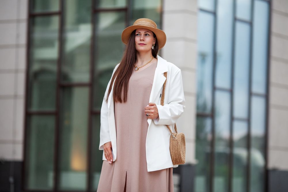 A woman in a beige dress and white blazer stands outdoors, wearing a hat and holding a woven bag, showcasing stylish outfit options.