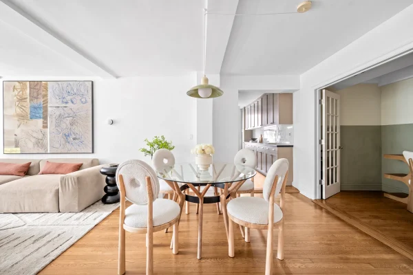 Modern dining area featuring a round glass table, stylish chairs, and a cozy sofa, with a backdrop of elegant artwork and a kitchen.