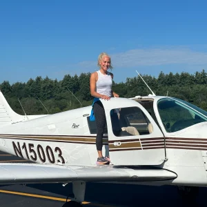 A woman stands on the wing of a small airplane, smiling proudly against a clear blue sky at an airport.