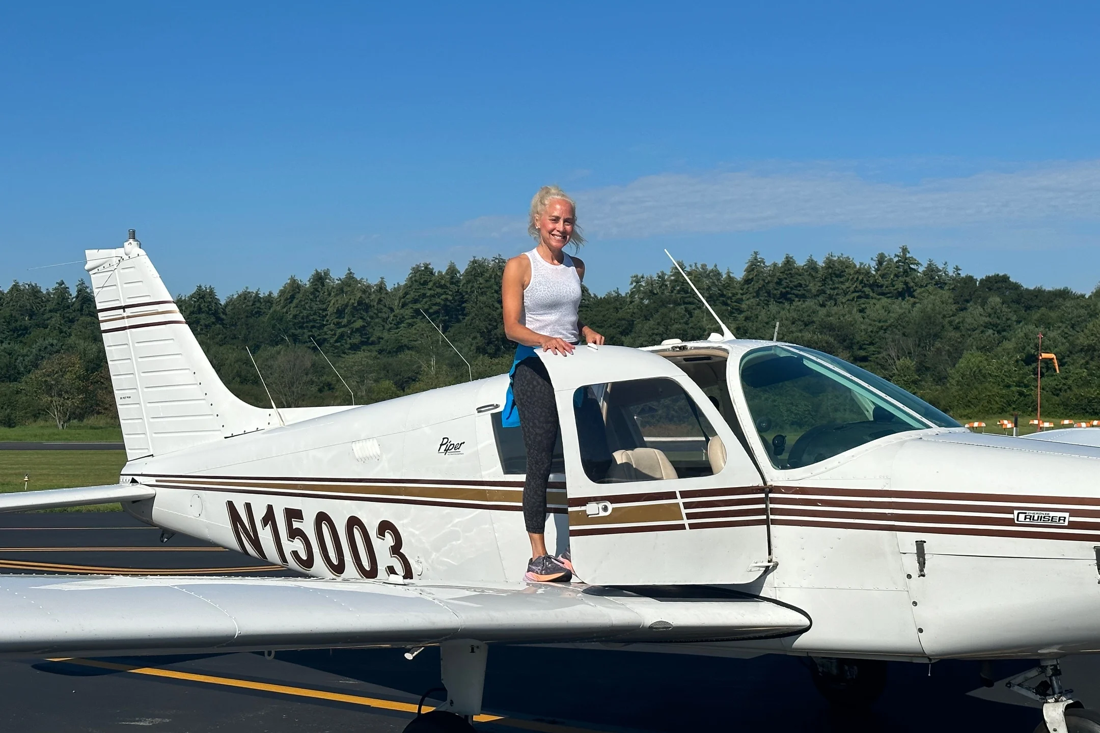 A woman stands on the wing of a small airplane, smiling proudly against a clear blue sky at an airport.