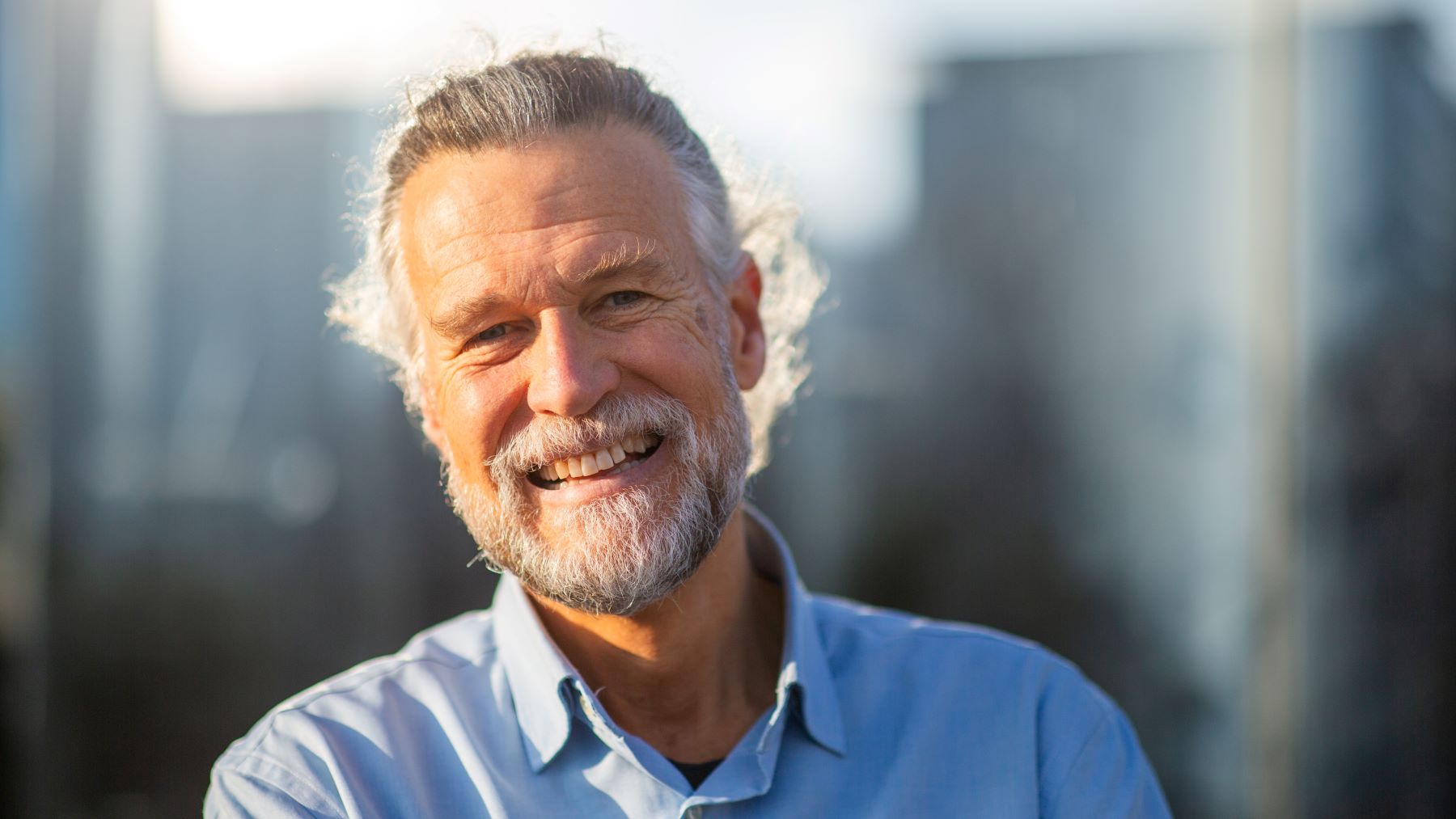 Smiling older man with a gray beard and long hair, wearing a blue shirt against a blurred background.