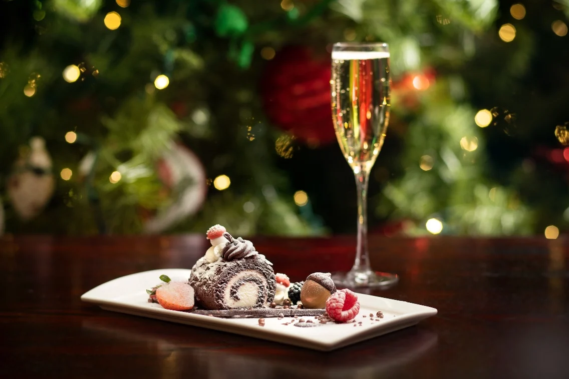 A festive yule log dessert with chocolate, cream, and berries, accompanied by a glass of champagne against a holiday backdrop.