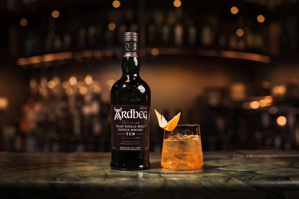 Ardbeg Ten whisky bottle beside an Old Fashioned cocktail garnished with orange peel on a dark bar countertop.