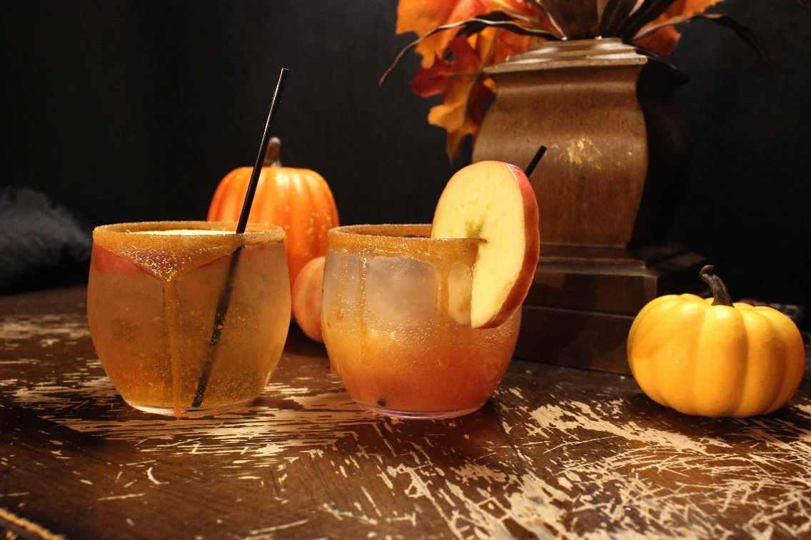 Two decorated glasses with apple-themed drinks sit on a rustic table, surrounded by decorative pumpkins and fall foliage.