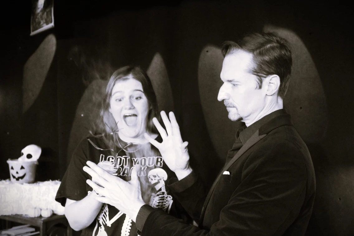 A magician performs a trick while a surprised audience member reacts in excitement, surrounded by Halloween decorations.