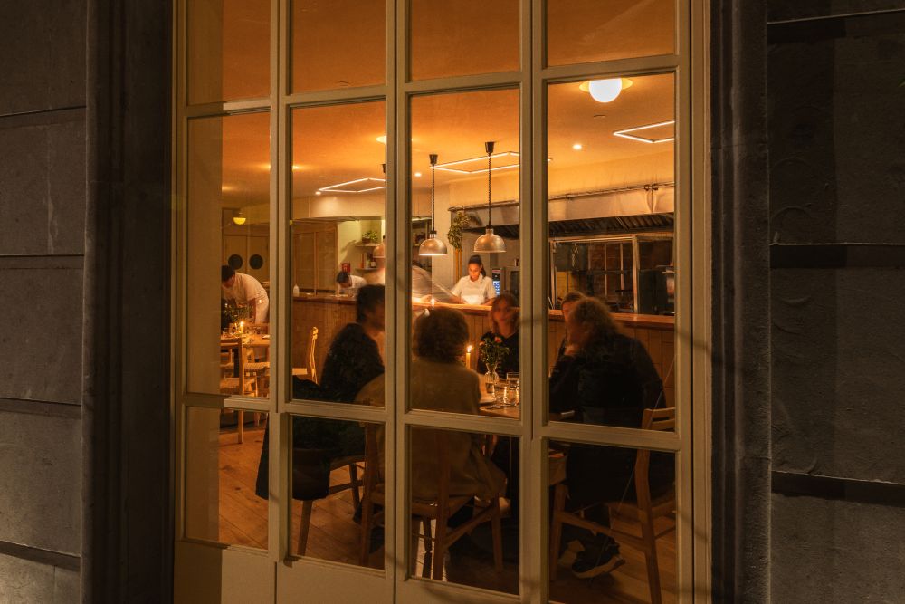 Warmly lit restaurant interior seen through a large window, with diners at a table and a chef visible in the background.