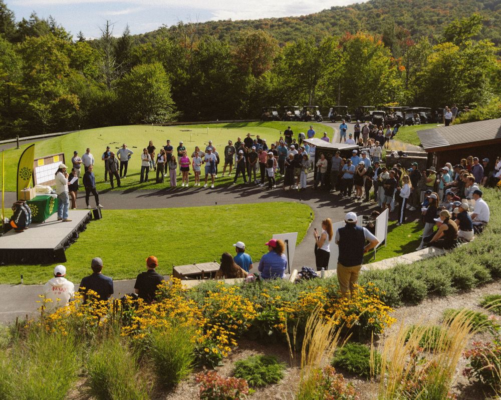 A crowd gathers around a stage at a golf event, enjoying speeches and the scenery in a vibrant outdoor setting.