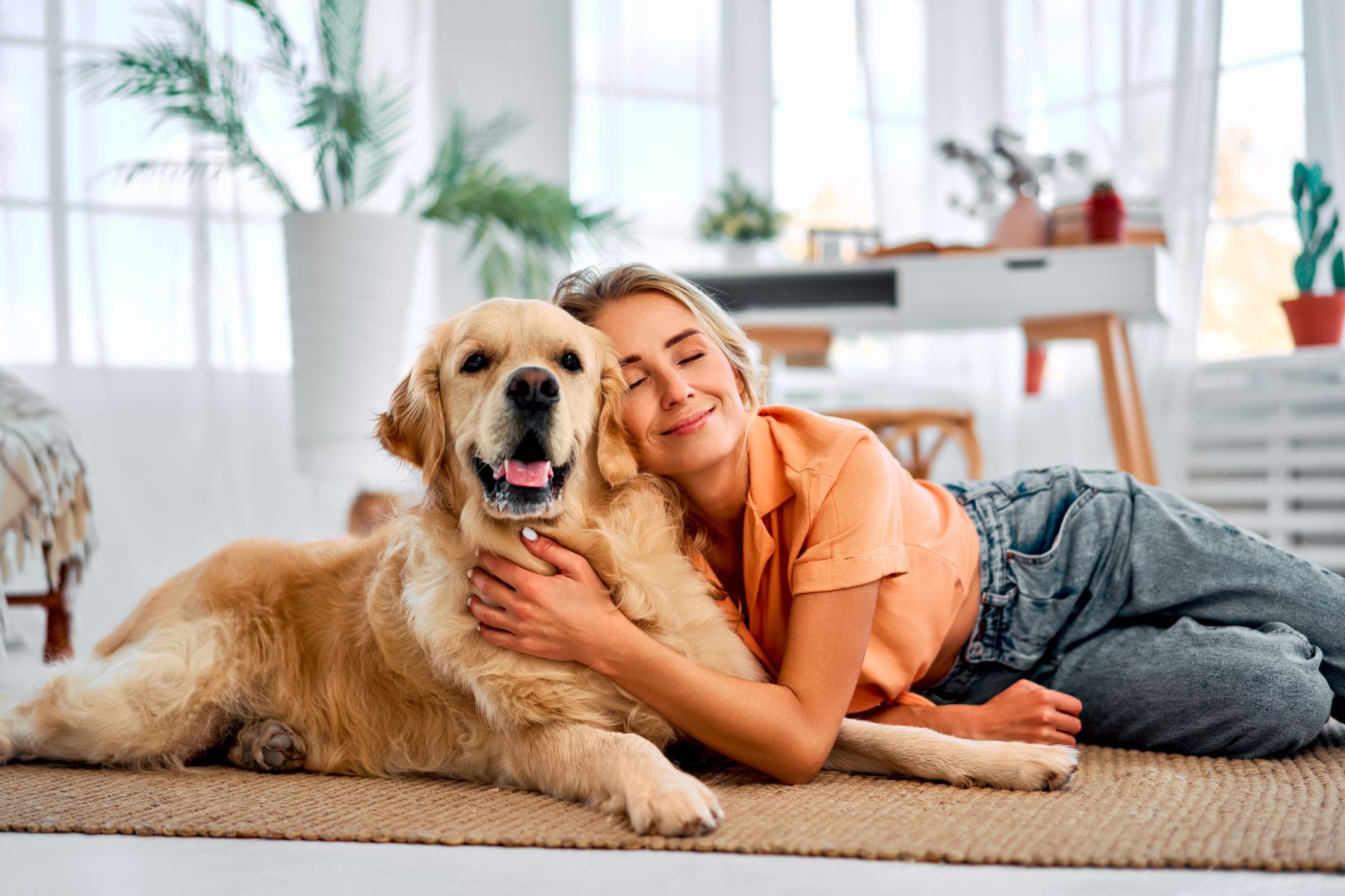 Woman happily cuddles with a golden retriever on a cozy rug in a bright, airy room, showcasing their bond.