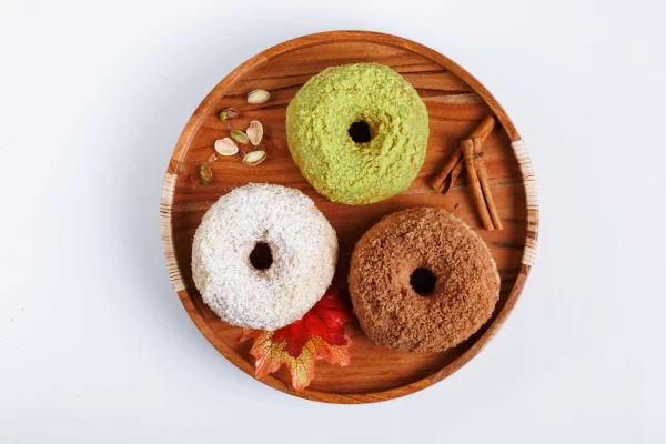 Three seasonal donuts in a wooden bowl, featuring flavors like pistachio, coconut, and cinnamon, garnished with leaves and nuts.