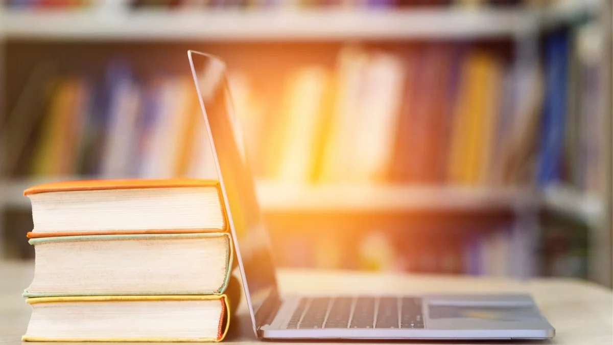 A stack of three books next to an open laptop on a table, set against a blurred background of bookshelves.