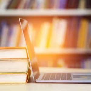 A stack of three books next to an open laptop on a table, set against a blurred background of bookshelves.