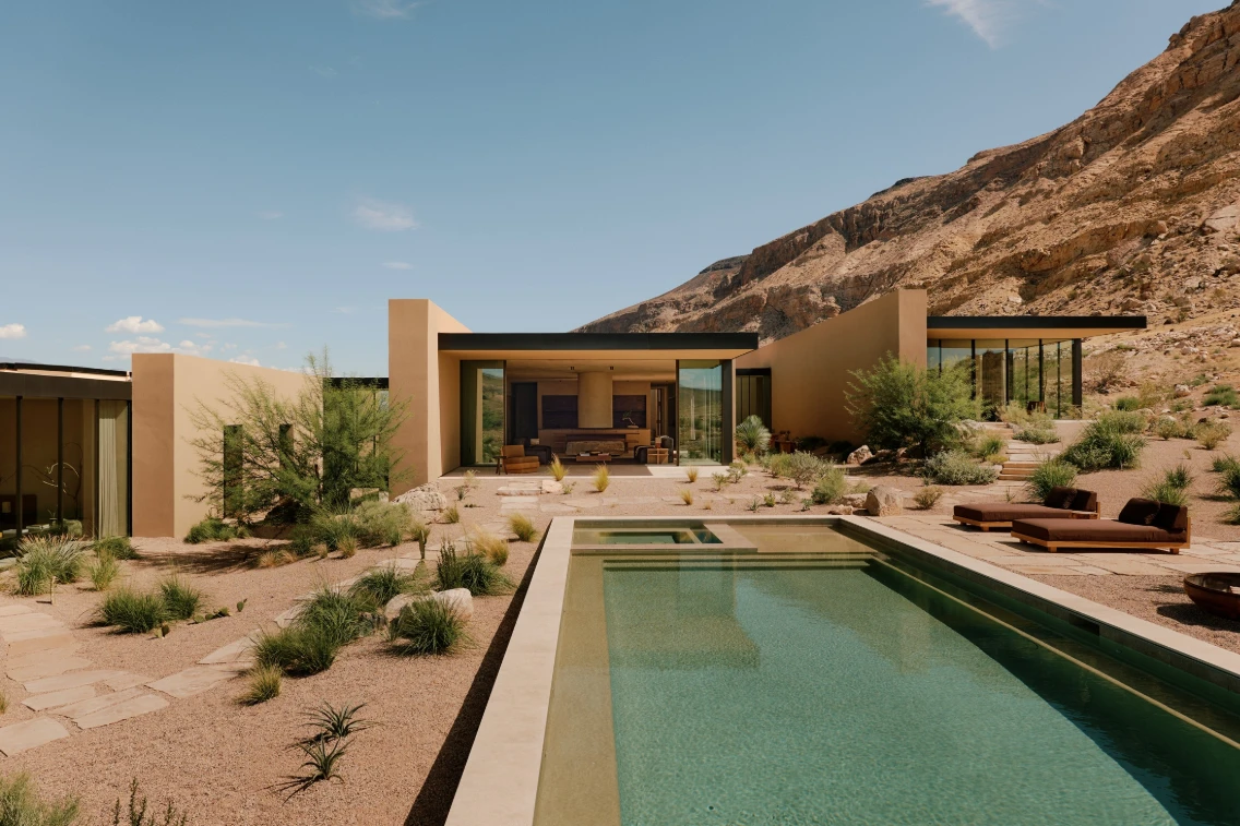 Modern desert retreat designed to blend into the landscape, featuring a pool and natural landscaping against mountain backdrop.