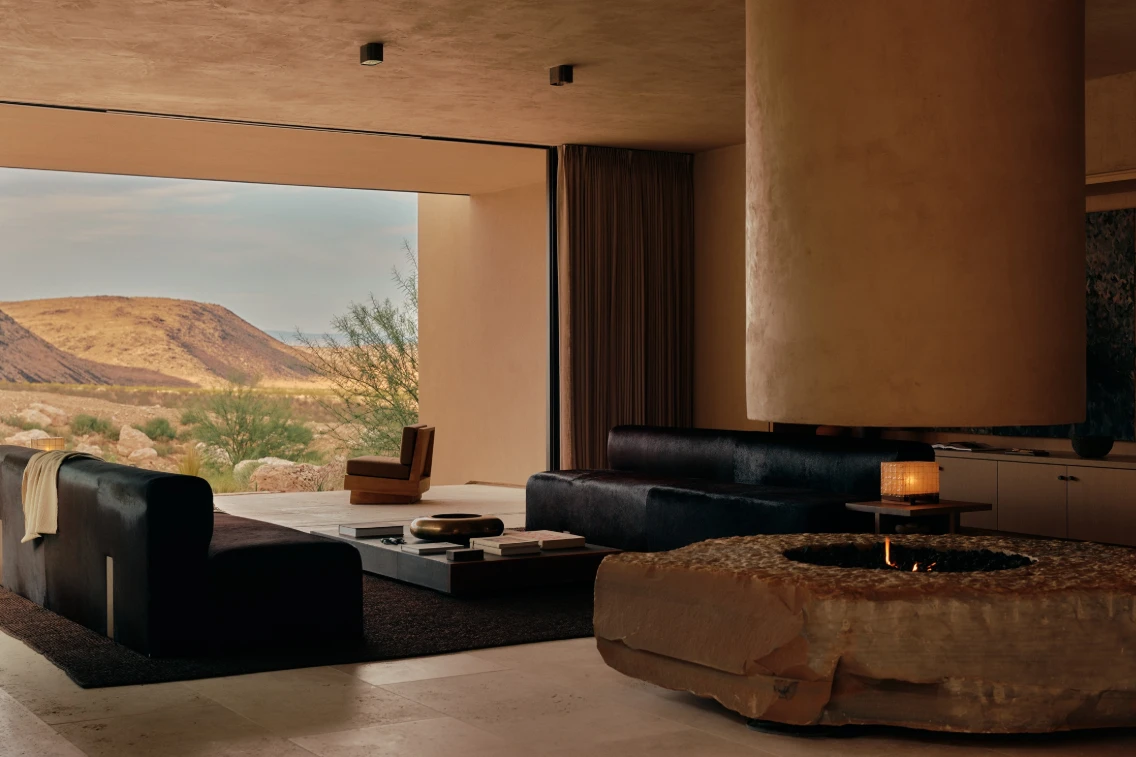 Modern living room featuring large black sofas, a unique stone coffee table, and expansive desert views through open windows.