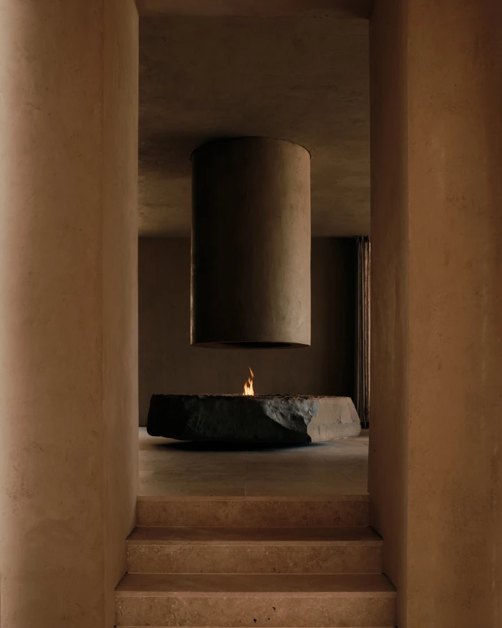 A modern interior featuring a suspended cylindrical feature above a dark stone fireplace in a minimalistic, earthy setting.