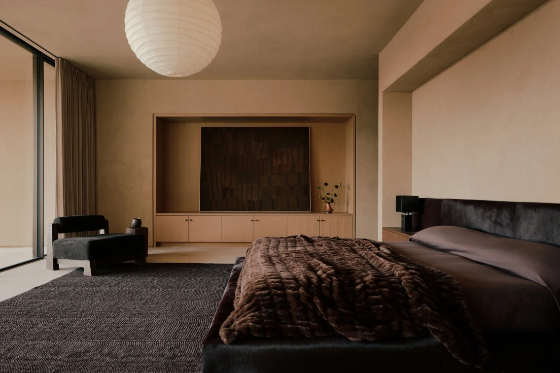 A modern bedroom featuring earthy tones, a plush bed with a cozy blanket, and minimalist furniture with large windows.