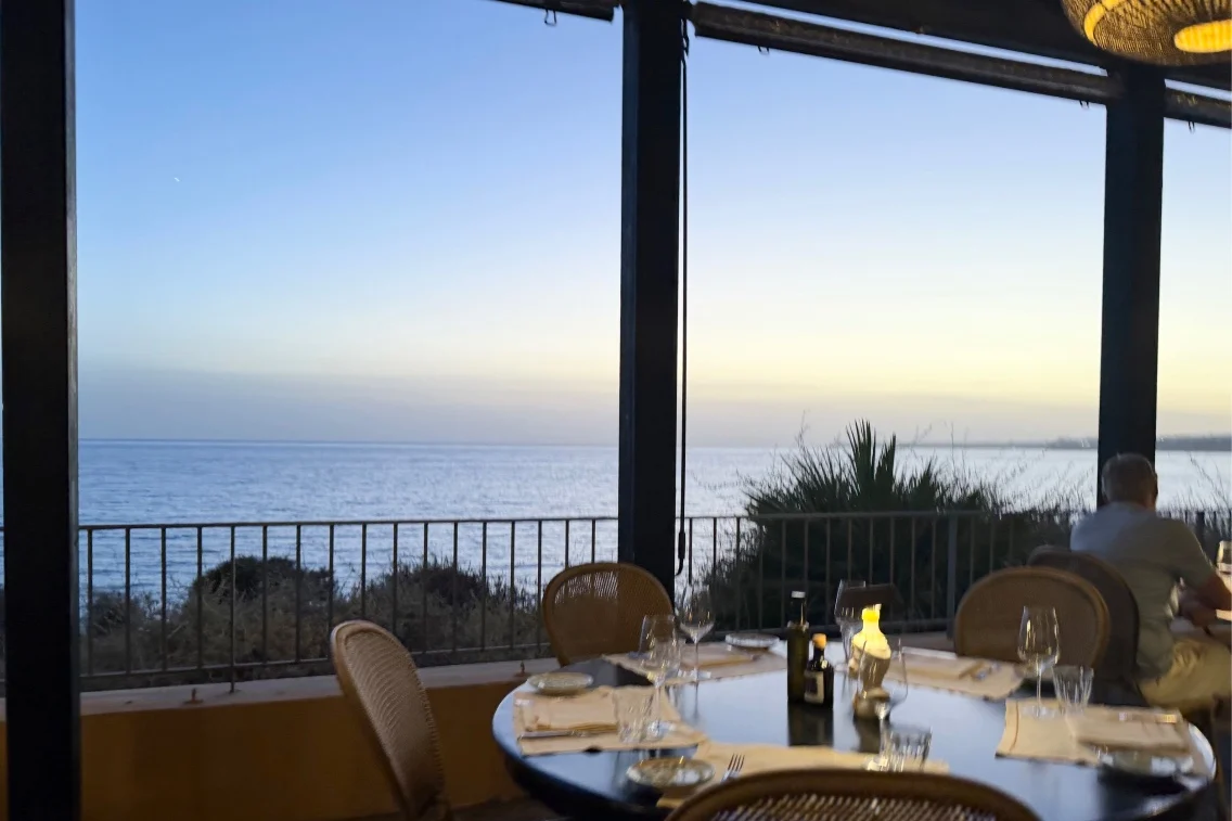 Dining area with a table set for a meal, overlooking the sea at sunset, with a beachy ambiance at Verdura Resort.
