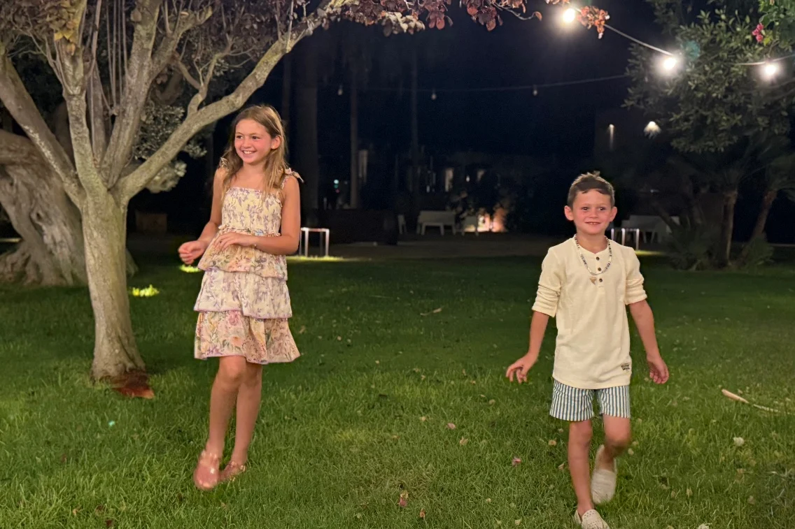 Two children playfully walk on grass at night, surrounded by gentle lighting and a serene atmosphere at the resort.