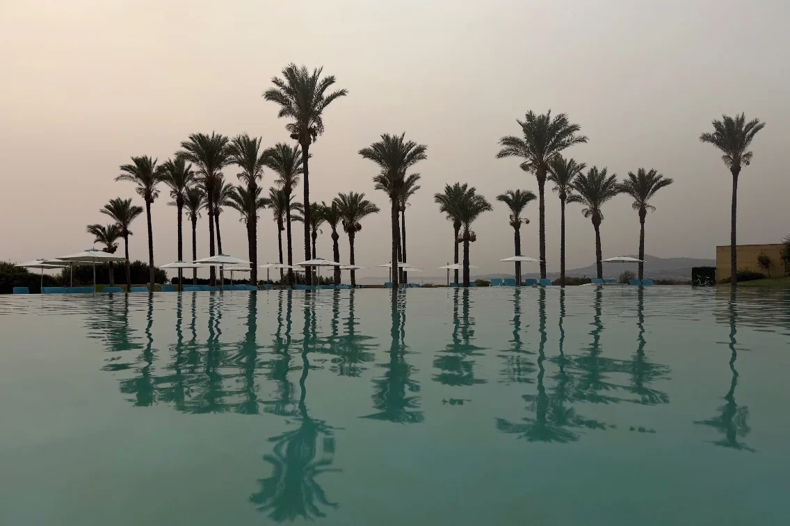 Still water reflecting palm trees and sun loungers at Rocco Forte's Verdura Resort during a hazy sunset.