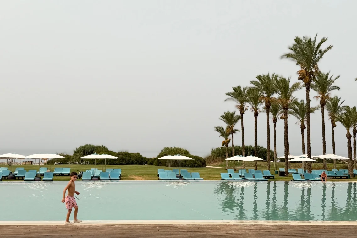 A child walks along the edge of a serene pool surrounded by palm trees and lounge chairs at Rocco Forte’s Verdura Resort.
