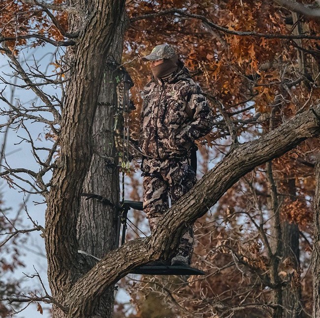 Hiker in camouflage apparel stands on a tree branch, showcasing style and functionality in hunting clothing.