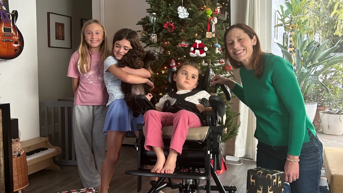Four children and a woman smile in front of a decorated Christmas tree with gifts around them, celebrating the holiday spirit.