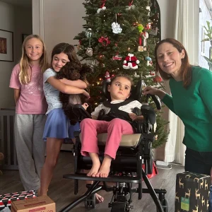 Four children and a woman smile in front of a decorated Christmas tree with gifts around them, celebrating the holiday spirit.