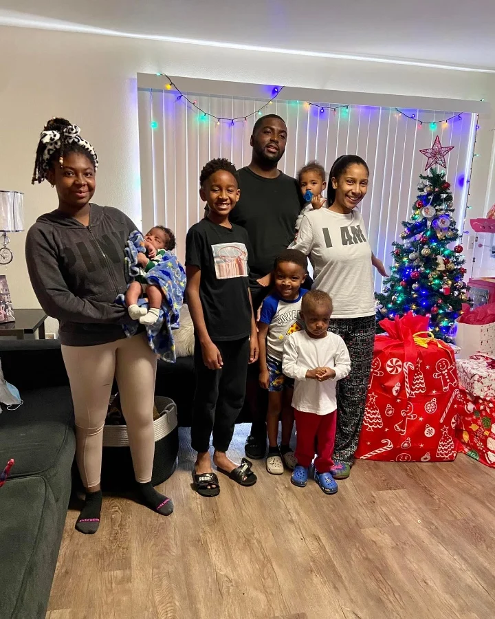 A smiling family stands together in a decorated living room with a Christmas tree and gifts.