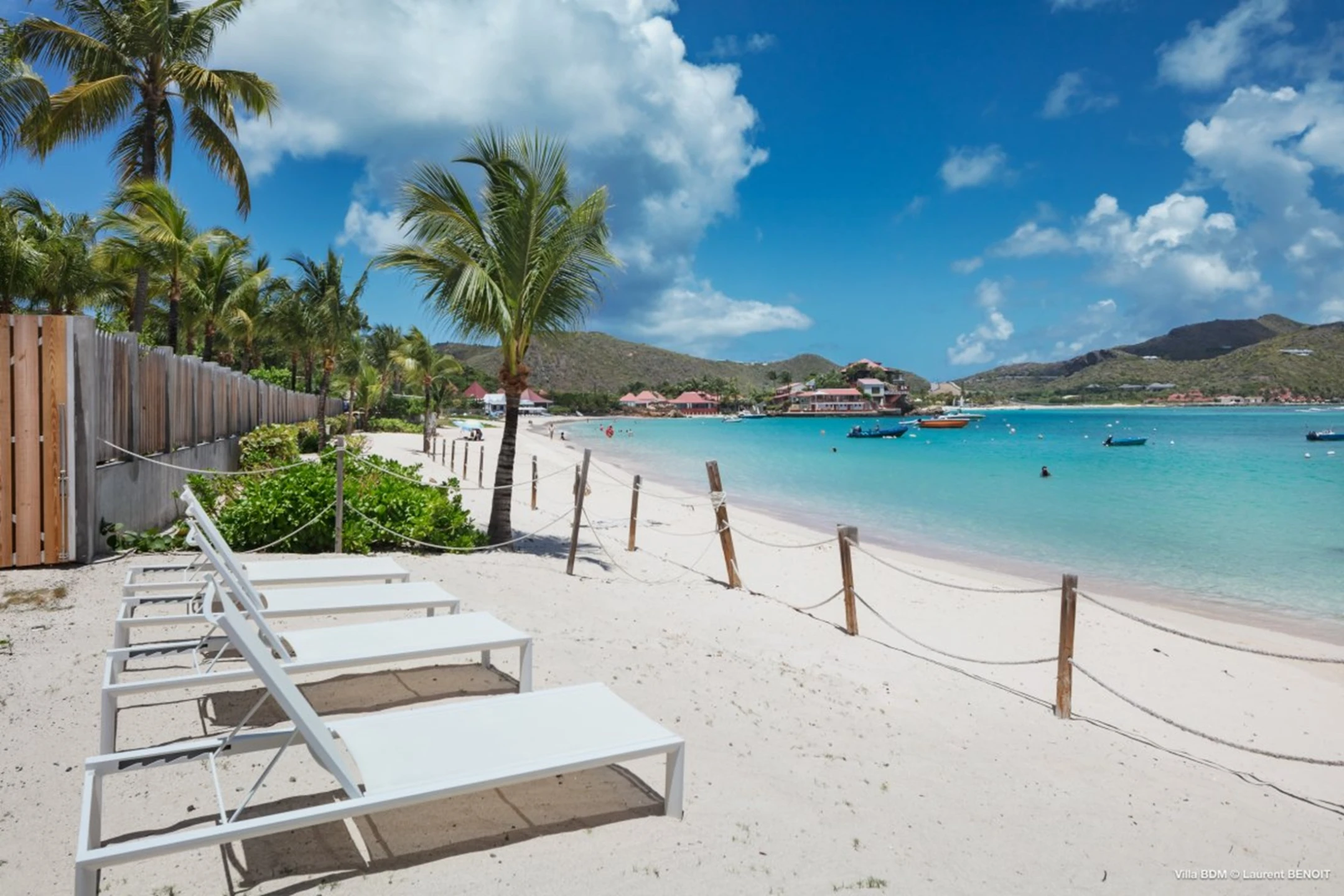 Lounge chairs line a sandy beach with clear turquoise water and palm trees in the background, capturing the essence of luxury in St. Barts.