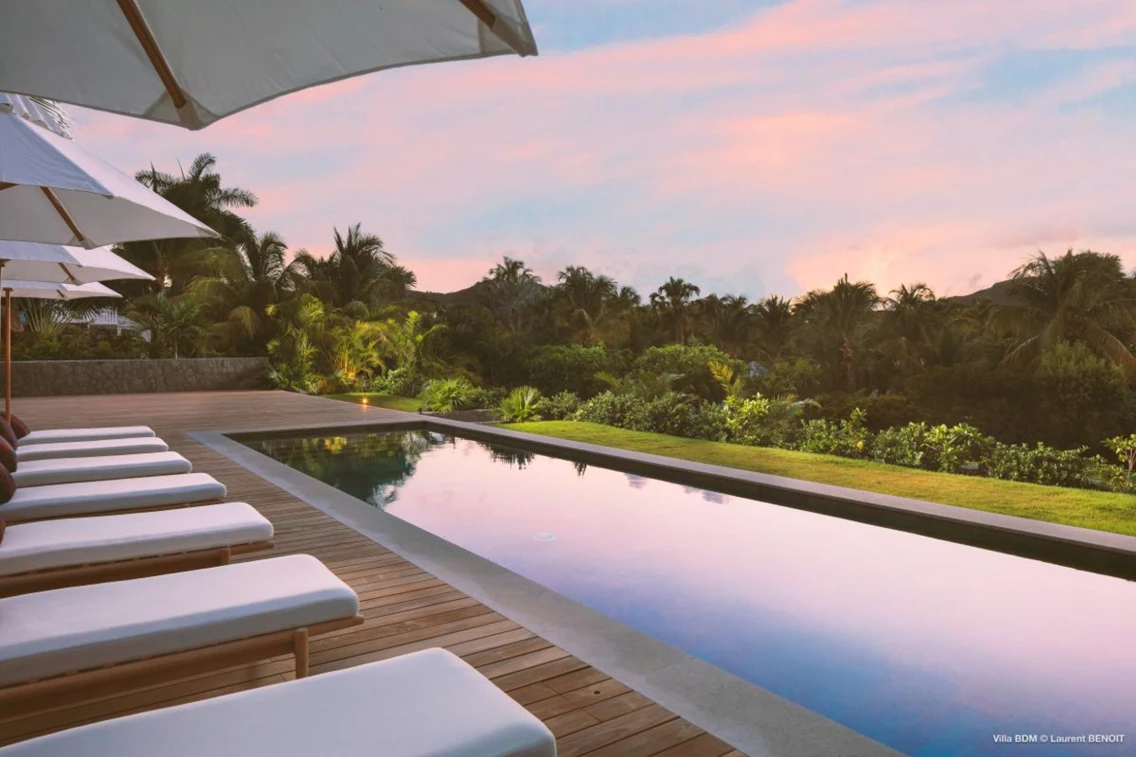 Luxurious villa exterior featuring a private pool, sun loungers, and tropical landscaping against a colorful sunset sky.