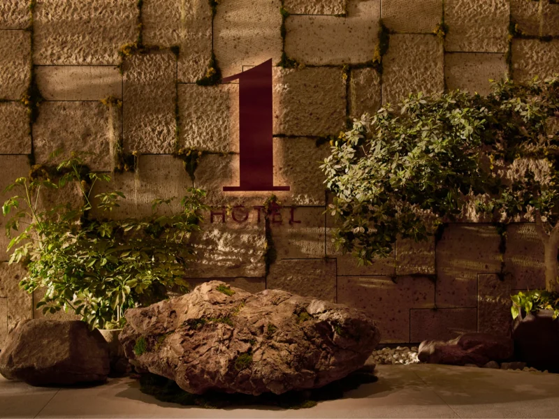 The sign for 1 Hotel Tokyo is displayed on a textured stone wall, flanked by rocks and greenery, creating a natural atmosphere.