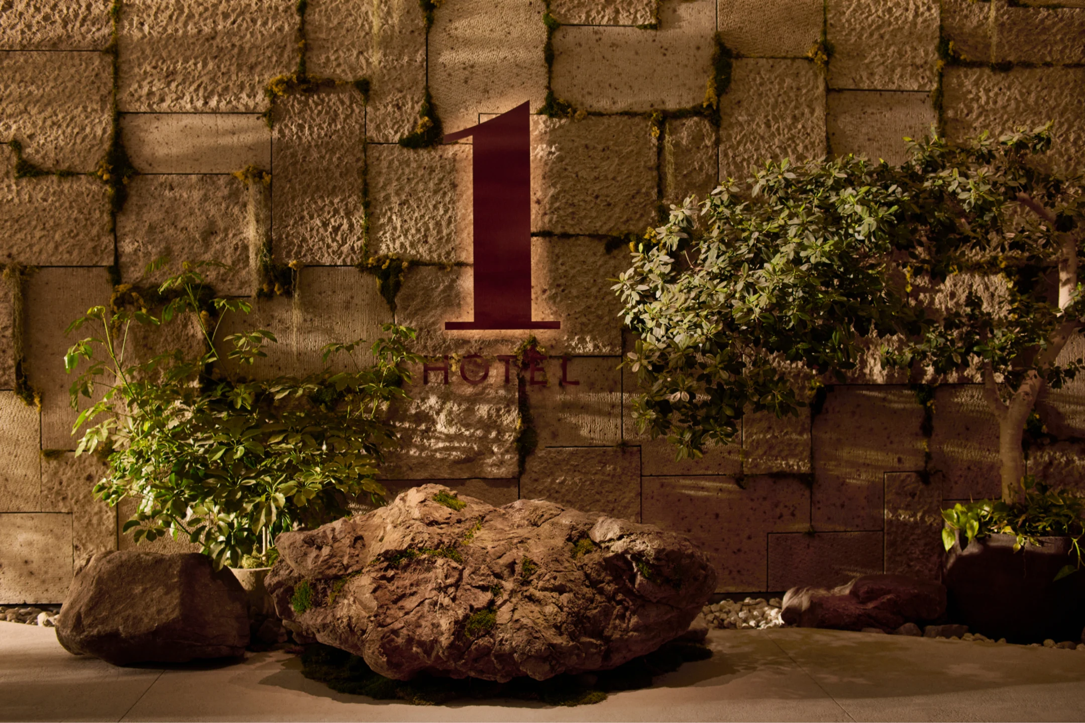 The sign for 1 Hotel Tokyo is displayed on a textured stone wall, flanked by rocks and greenery, creating a natural atmosphere.